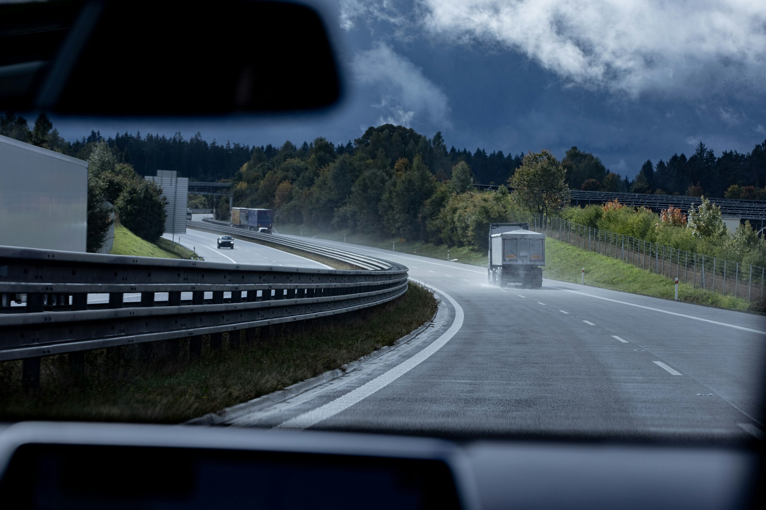 View from inside a car driving on a wet highway surrounded by trees, with a truck ahead and dark storm clouds looming in the sky, creating a moody and tense atmosphere.