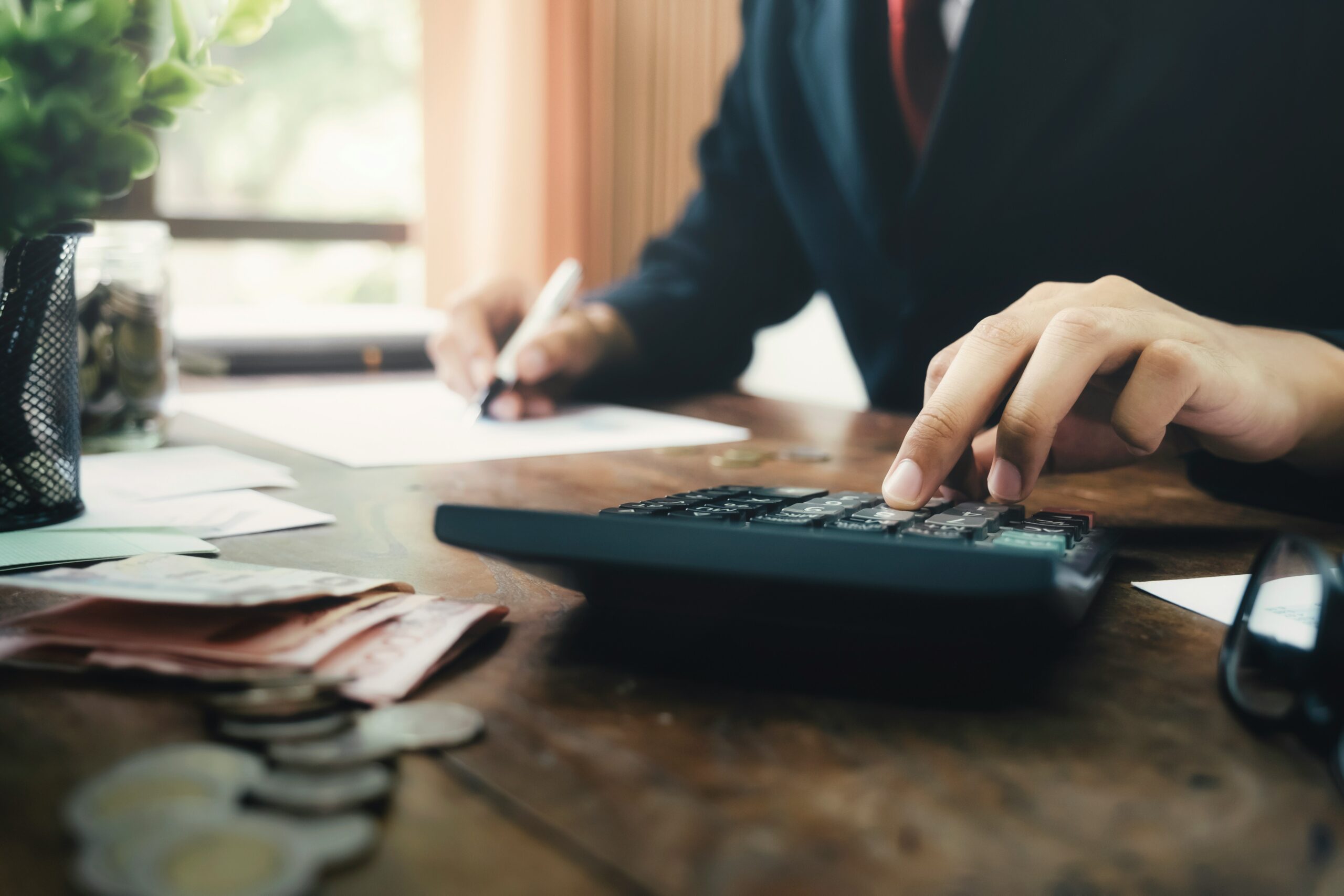 A person in a suit works at a desk, using a calculator with one hand while writing on documents with the other, surrounded by papers, coins, and banknotes in a softly lit office setting.