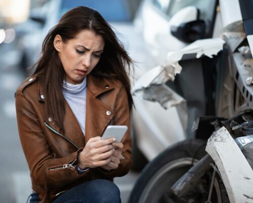 A distressed woman kneels beside a badly damaged white car, looking down at her phone with concern as she appears to document or report the accident on a busy street.