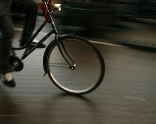 A blurred, motion-filled close-up of a person riding a bicycle, capturing the front wheel and pedaling legs as the bike moves quickly along a paved street.