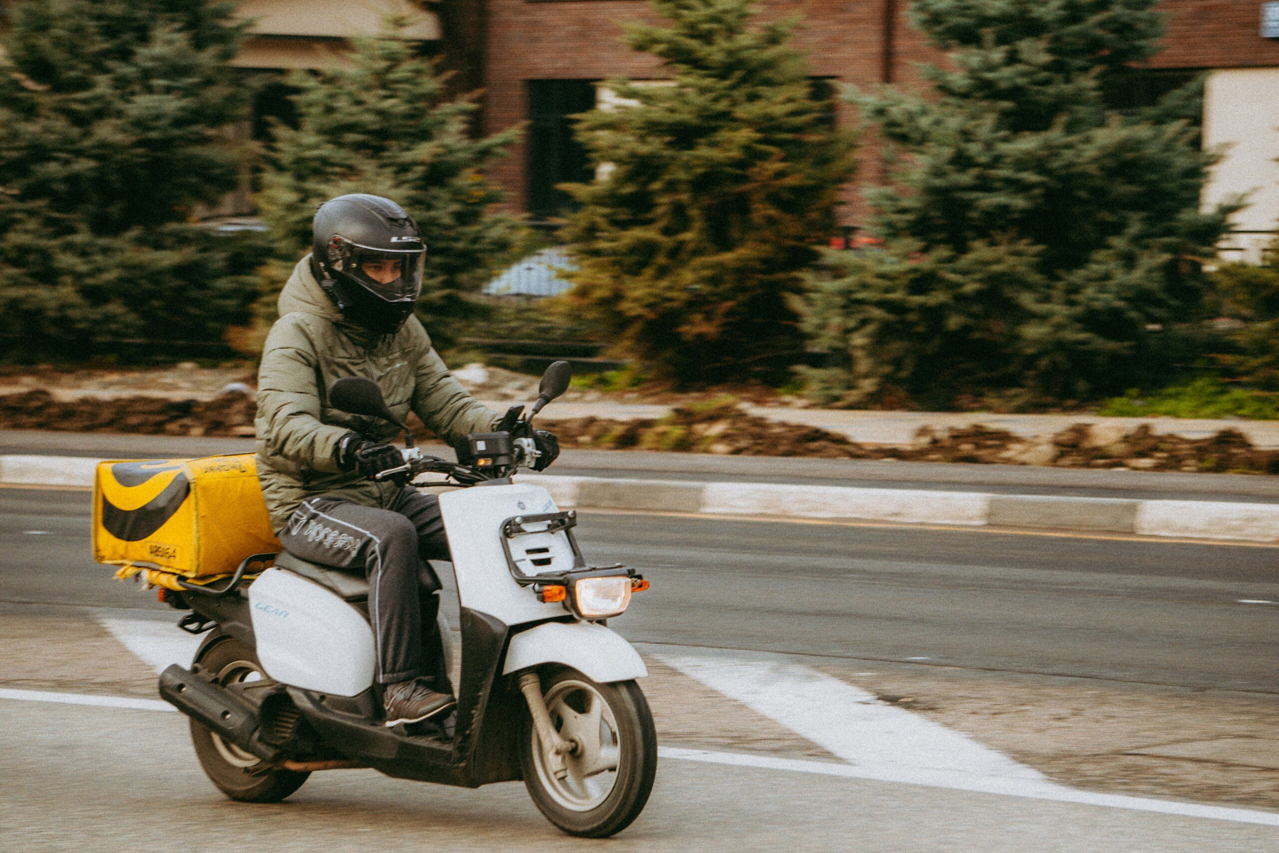 A delivery rider wearing a helmet and padded jacket drives a white scooter with a yellow cargo box attached to the back, traveling along a city street lined with evergreens.