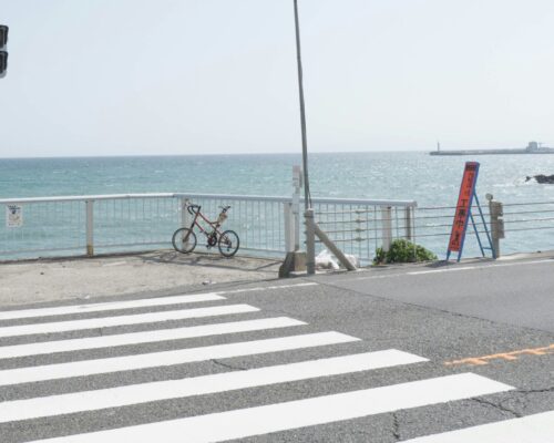 Crosswalk leading to a seaside view with a bicycle parked near a railing, traffic signs, and a red pedestrian light under a clear blue sky.