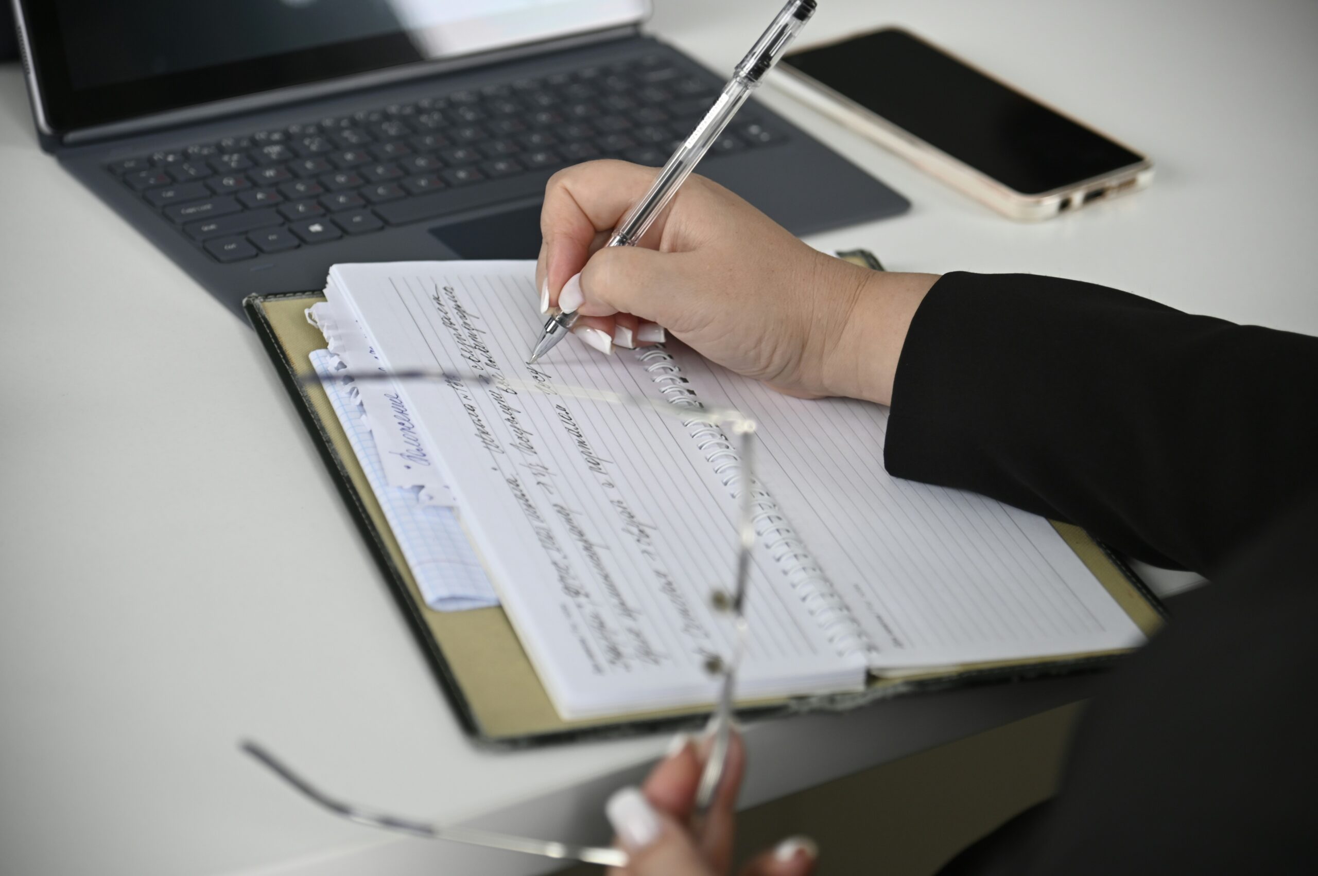 A close-up view of a person’s hand writing notes with a silver pen in a lined spiral notebook resting on a desk, with handwritten text visible across the pages; a pair of eyeglasses is held loosely in the other hand, while a laptop keyboard and a smartphone lie slightly out of focus in the background, suggesting a work or study setting.
