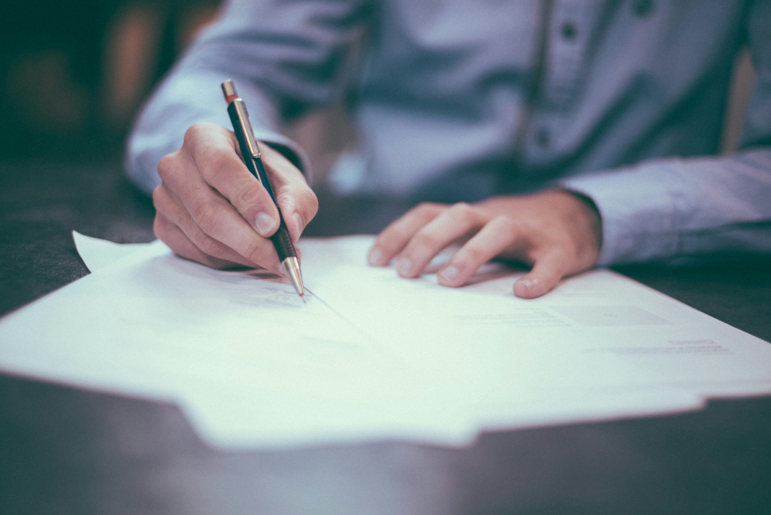 A close-up of a person’s hands as they write on a stack of documents with a pen, resting on a desk with papers spread out.