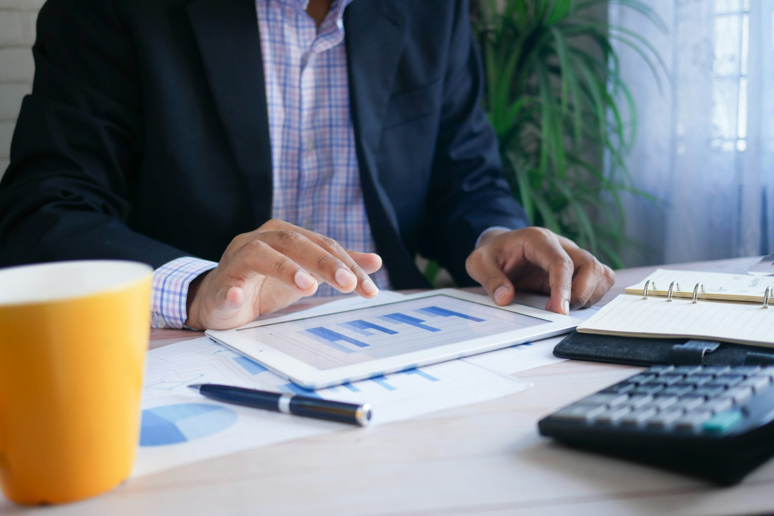 Business professional reviews bar chart data on a tablet at a desk with printed reports, a notebook, calculator, pen, and coffee mug in an office setting.