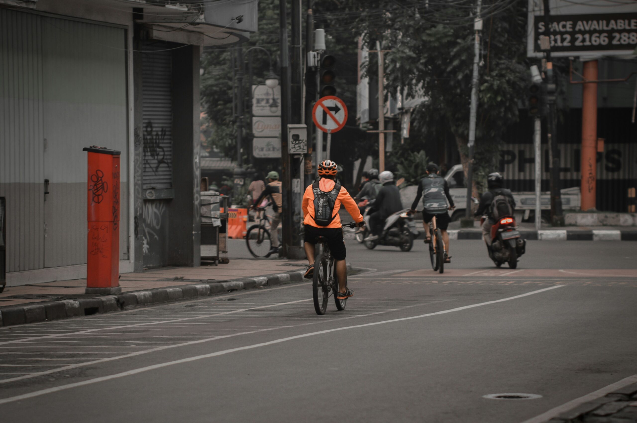 Urban street scene with a cyclist wearing an orange jacket and helmet riding in a designated bike lane toward an intersection, while several motorcycles and another cyclist travel ahead in mixed traffic, with buildings, trees, street signs, and a “no left turn” sign visible, creating a busy city environment focused on shared roadway use.