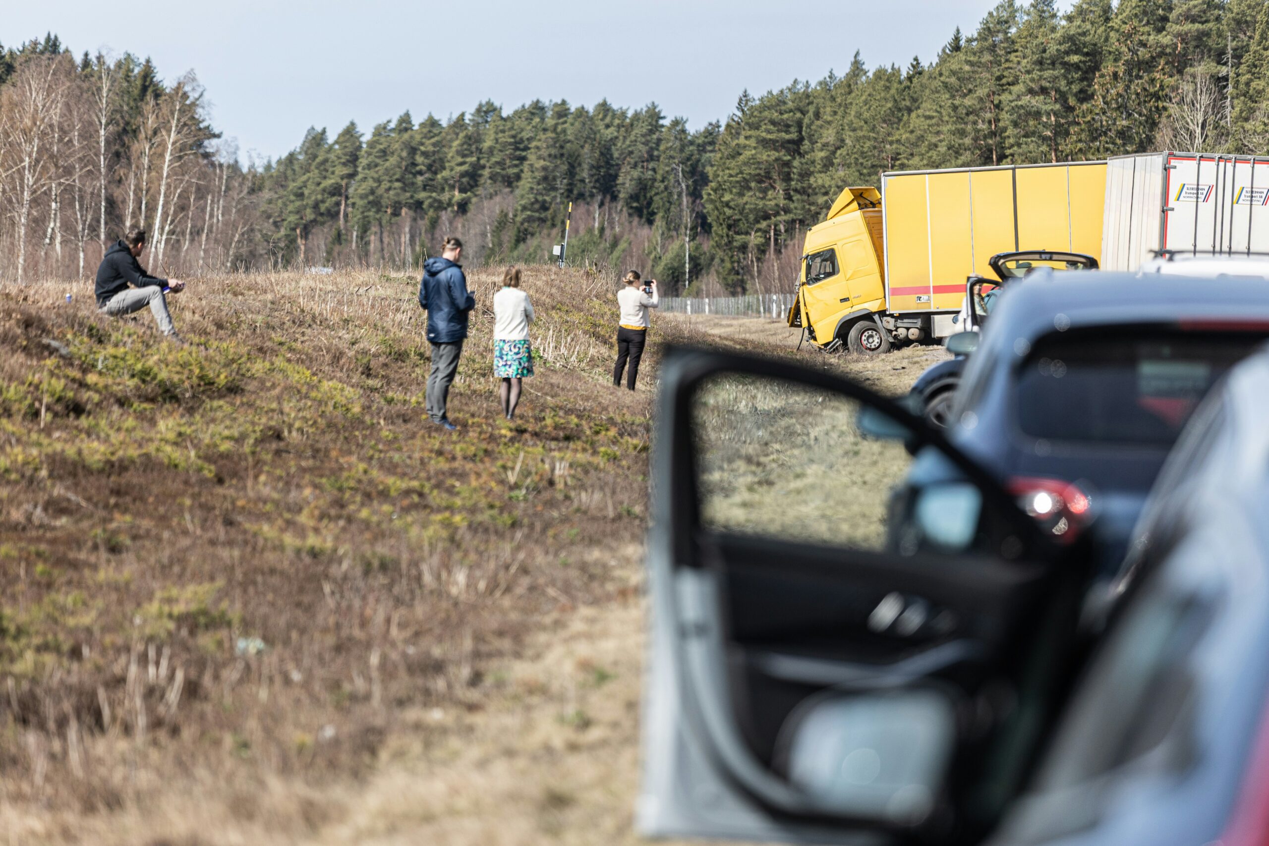 Roadside accident scene with a yellow box truck stopped off the road near a wooded area, several people standing on a grassy embankment observing or taking photos, and a parked car in the foreground with its door and side mirror visible.