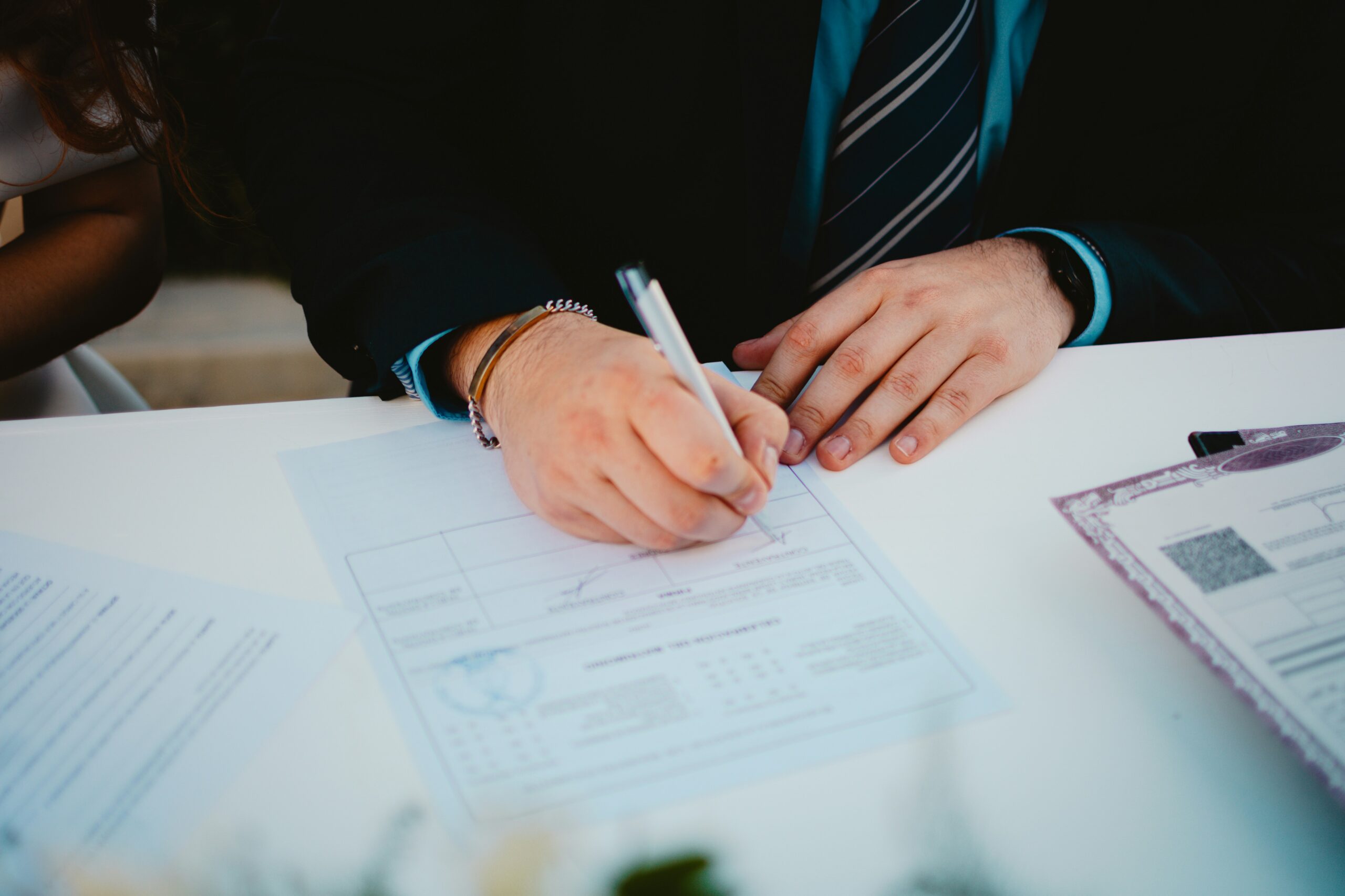 Close-up of a person in a suit and striped tie signing an official document on a white desk with a pen, with another person partially visible beside them and additional paperwork laid out nearby, suggesting a formal agreement or legal paperwork being completed.
