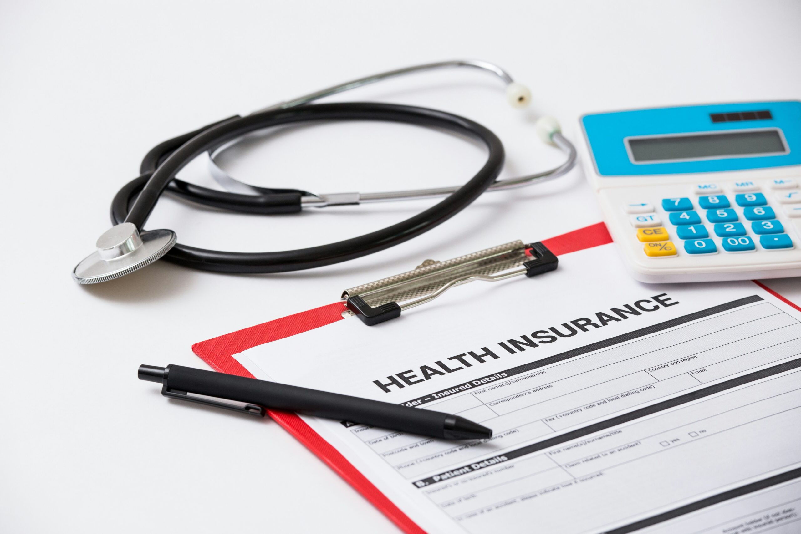 Stethoscope resting on a white surface beside a clipboard holding a health insurance form labeled “Health Insurance,” with a black pen placed across the document and a blue-and-white calculator positioned nearby, creating a clean, clinical arrangement associated with medical paperwork and insurance documentation.