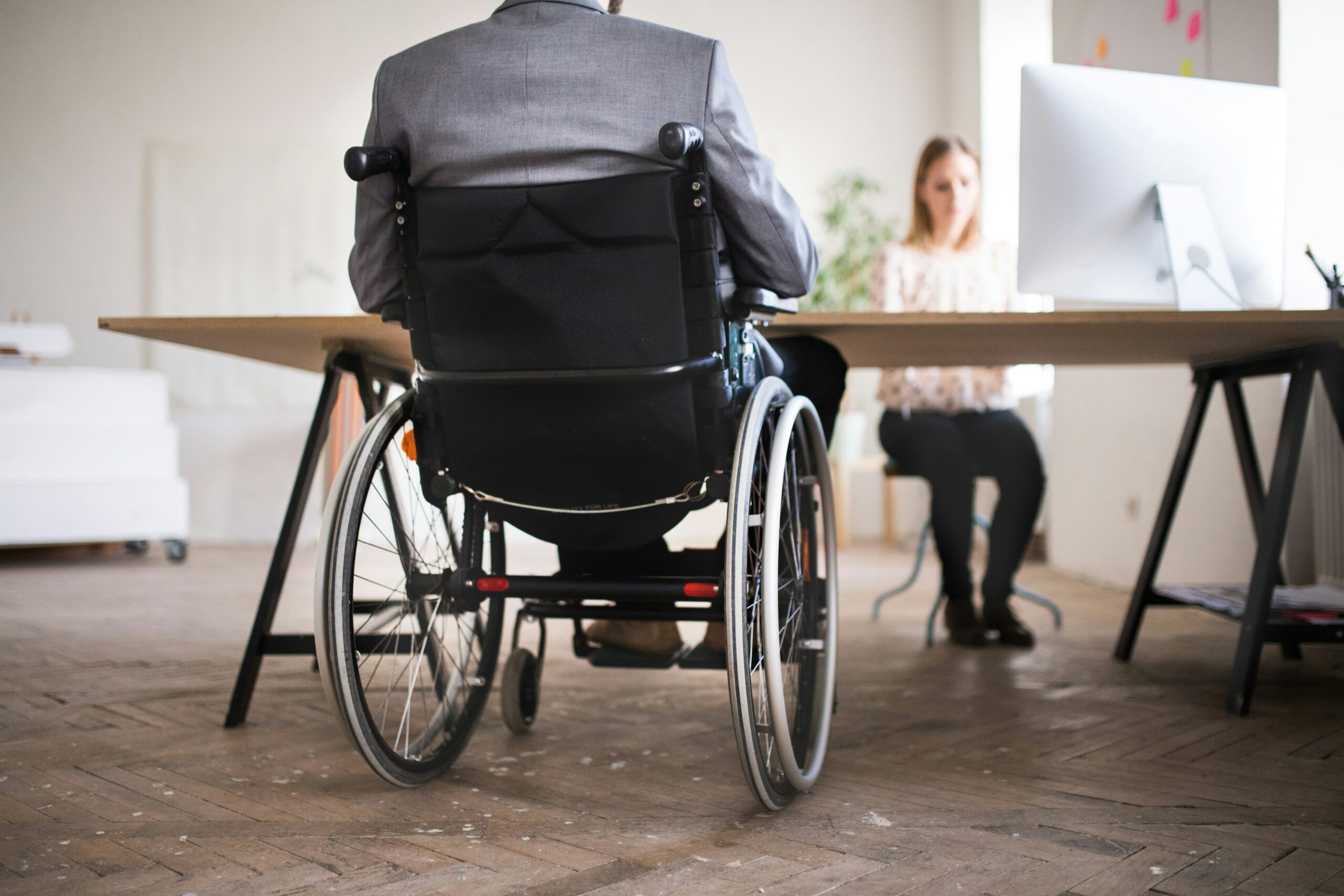 Person seated in a wheelchair wearing a gray suit jacket, viewed from behind at a wooden office desk, facing a woman seated across the table working at a large desktop computer in a bright, modern office space with wooden flooring, minimal furniture, and soft natural light, suggesting a formal meeting, consultation, or workplace discussion in an accessible professional environment.
