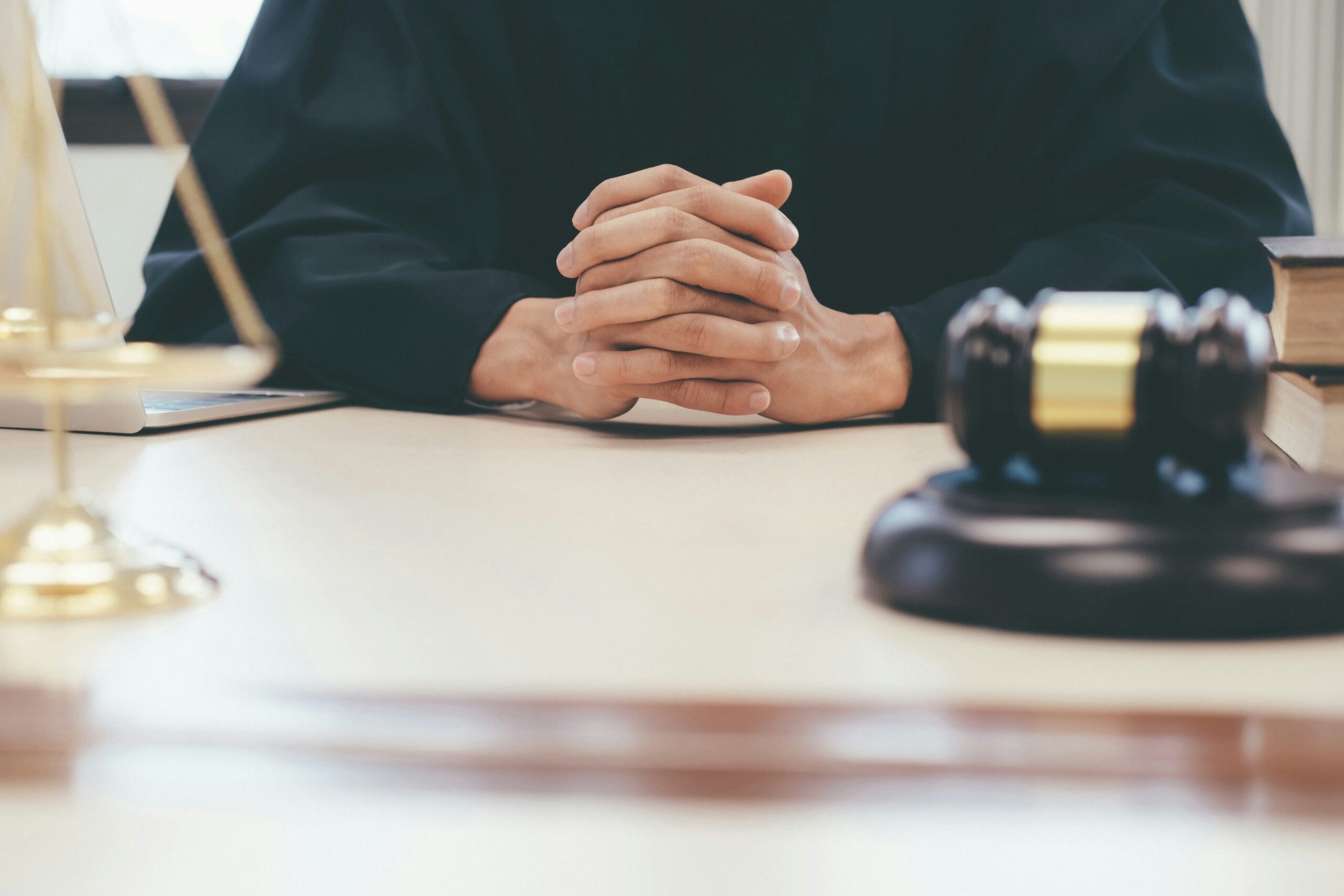 Close-up of a person in judicial robes seated at a desk with hands clasped together, a wooden judge’s gavel resting in the foreground, a brass balance scale partially visible to the side, and law books and a laptop on the desk, creating a formal courtroom or legal office setting that conveys authority and decision-making.