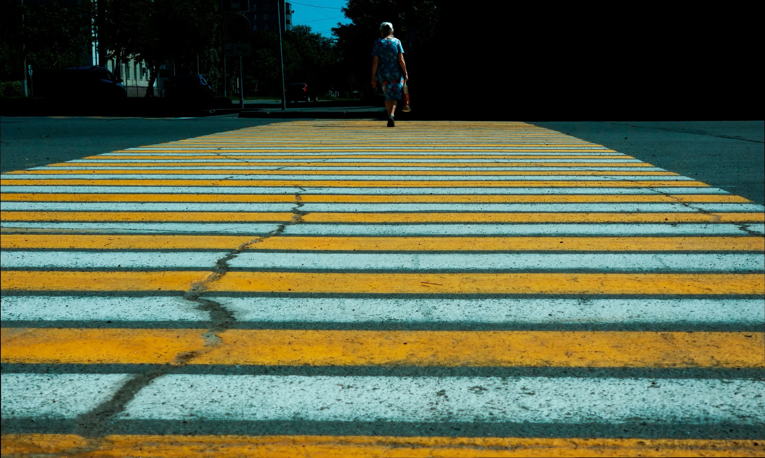 Low-angle view of a wide pedestrian crosswalk painted with alternating yellow and white stripes, cracked and weathered asphalt visible between the lines, with an elderly woman in a patterned dress walking away from the camera across the street, her figure small against the empty roadway, dark shaded surroundings and trees framing the scene, conveying distance and quiet movement through an urban intersection.