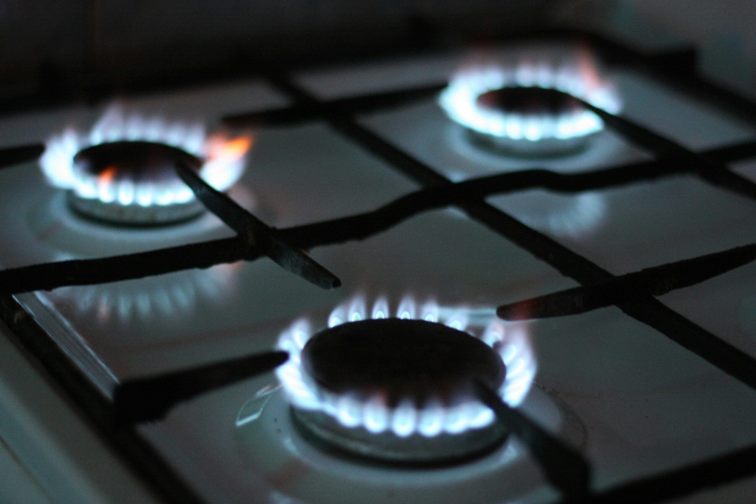 Close-up view of a modern gas stovetop with four burners ignited, each producing steady blue flames around dark circular burner caps, with black metal grates crossing over a glossy light-colored surface, creating a contrast between the cool blue fire, dark hardware, and reflective enamel finish.