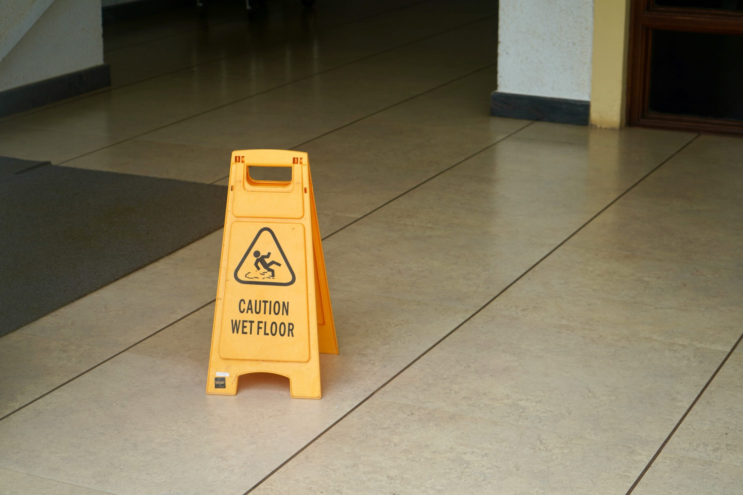 Yellow folding caution sign with a slipping figure icon and the words “Caution Wet Floor” printed on the front, placed on a light-colored tiled floor inside a building hallway, with clean tile grout lines, a dark floor mat to the left, and walls and door frames visible in the background, indicating a recently cleaned or potentially slippery indoor surface.