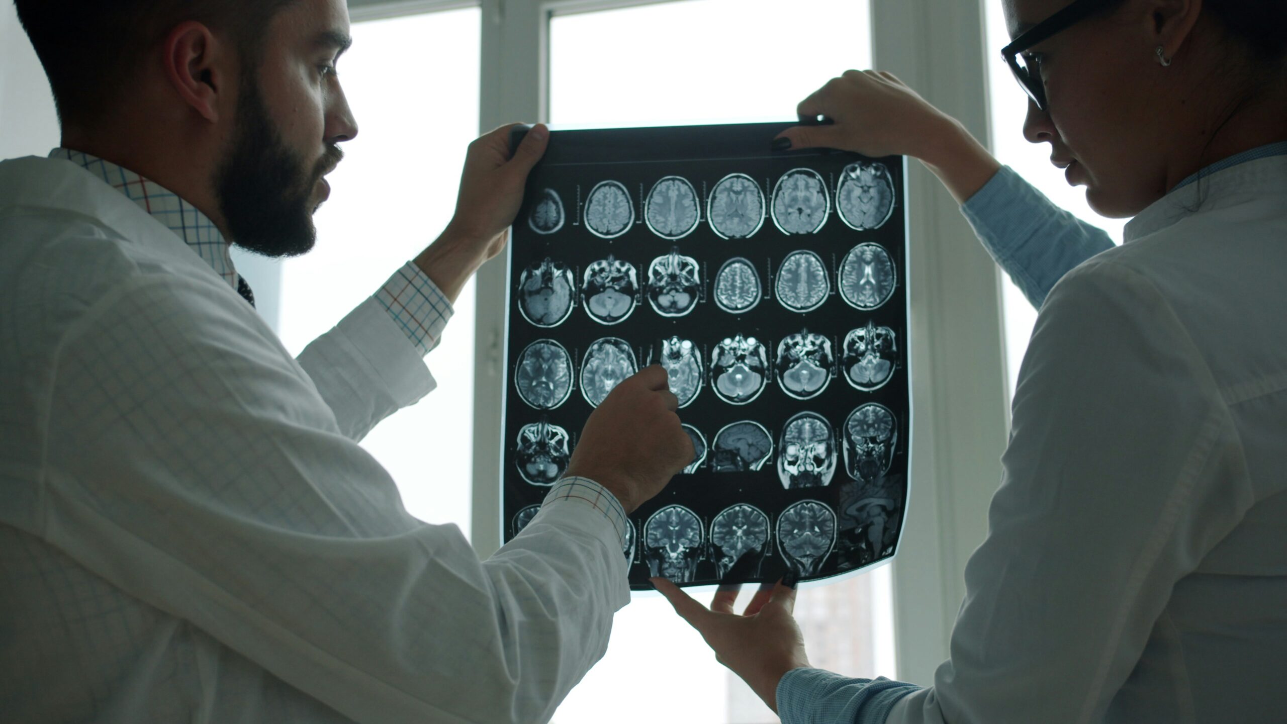 Two medical professionals in white coats examining a large sheet of brain scan images, holding it up against a bright window as one points to a specific area while they discuss the results together.