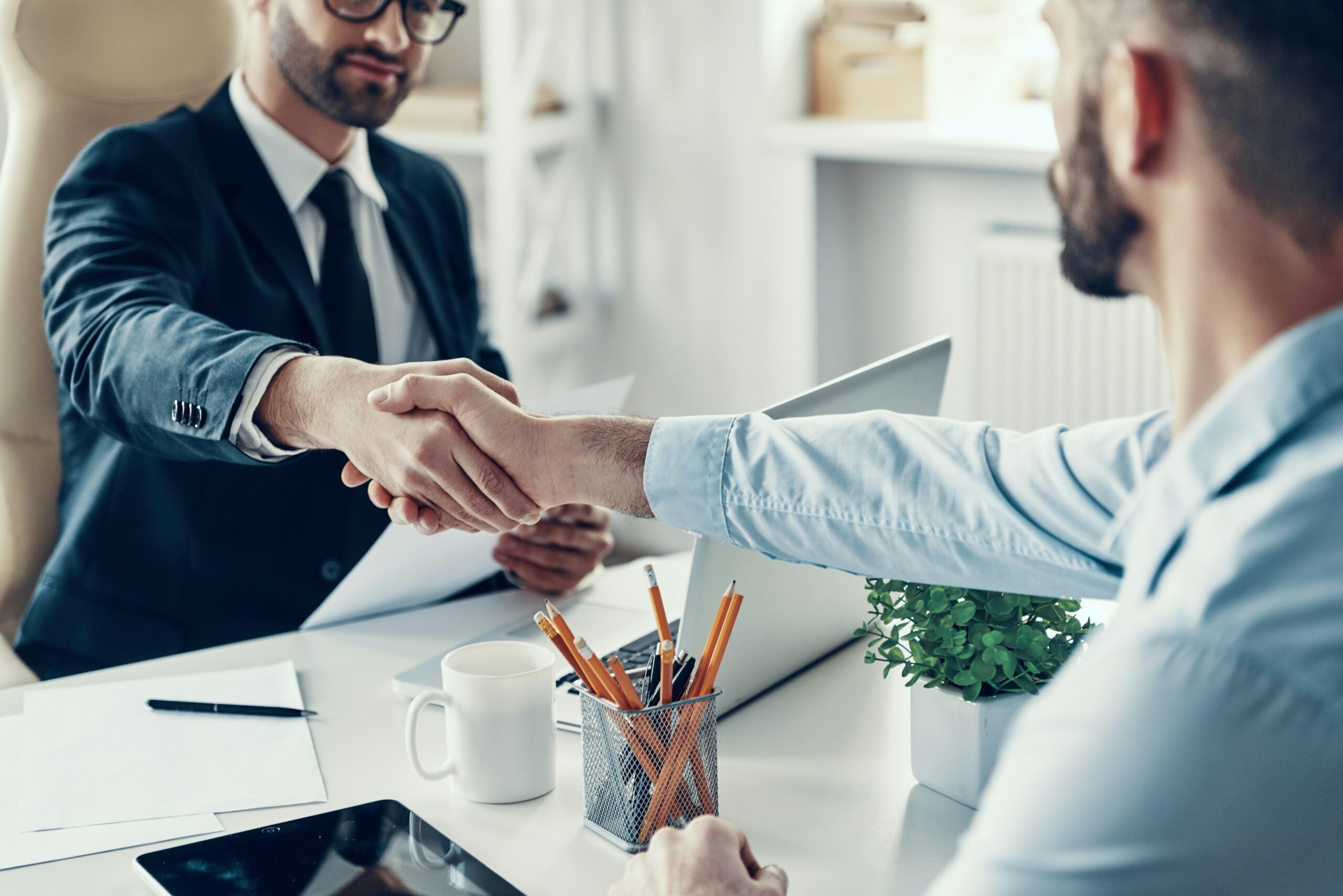 Two men shaking hands over a desk, symbolizing agreement or partnership in a professional setting.