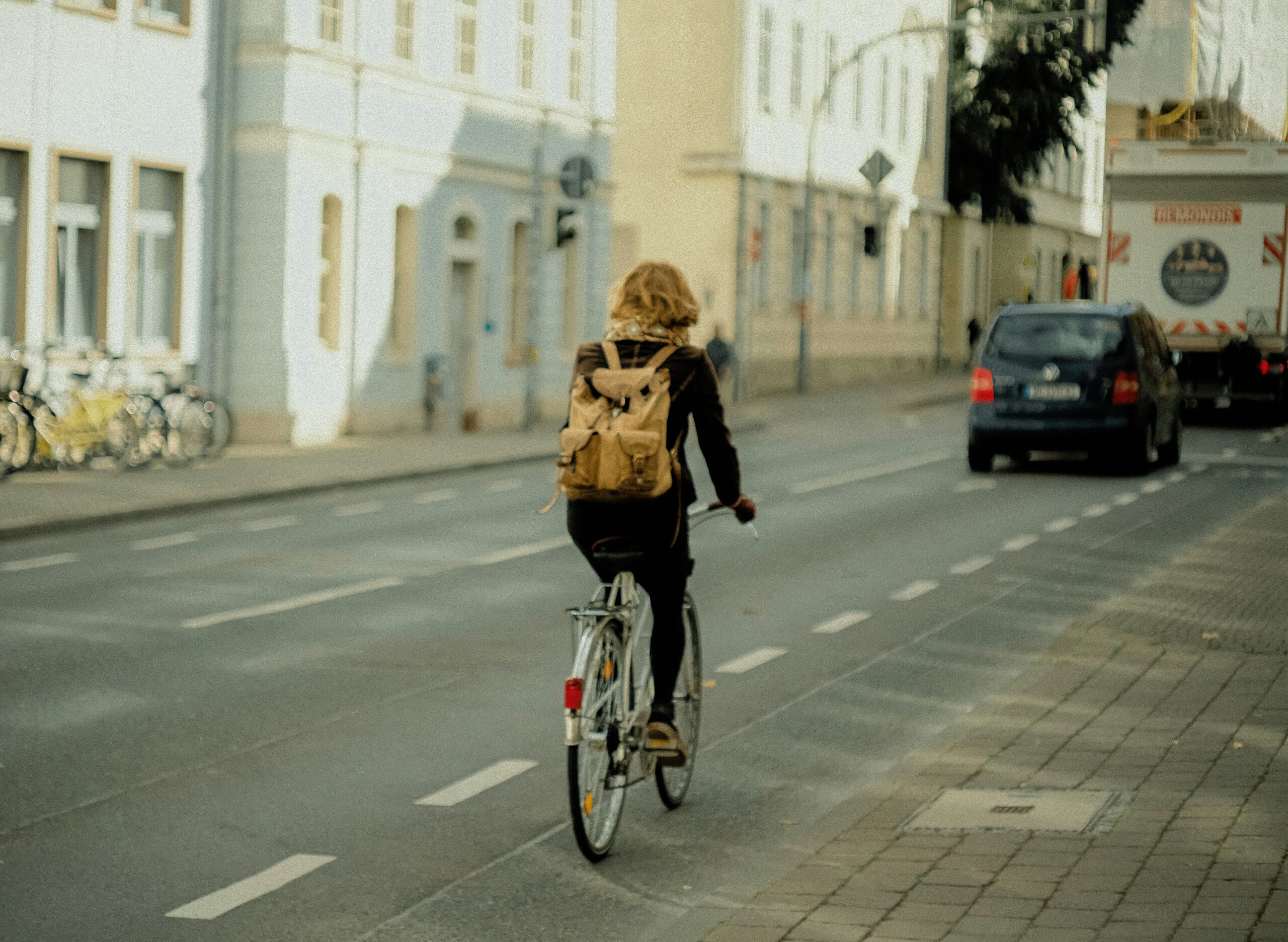 Person riding a bicycle along a city street while wearing a backpack, with cars driving ahead in the same lane.