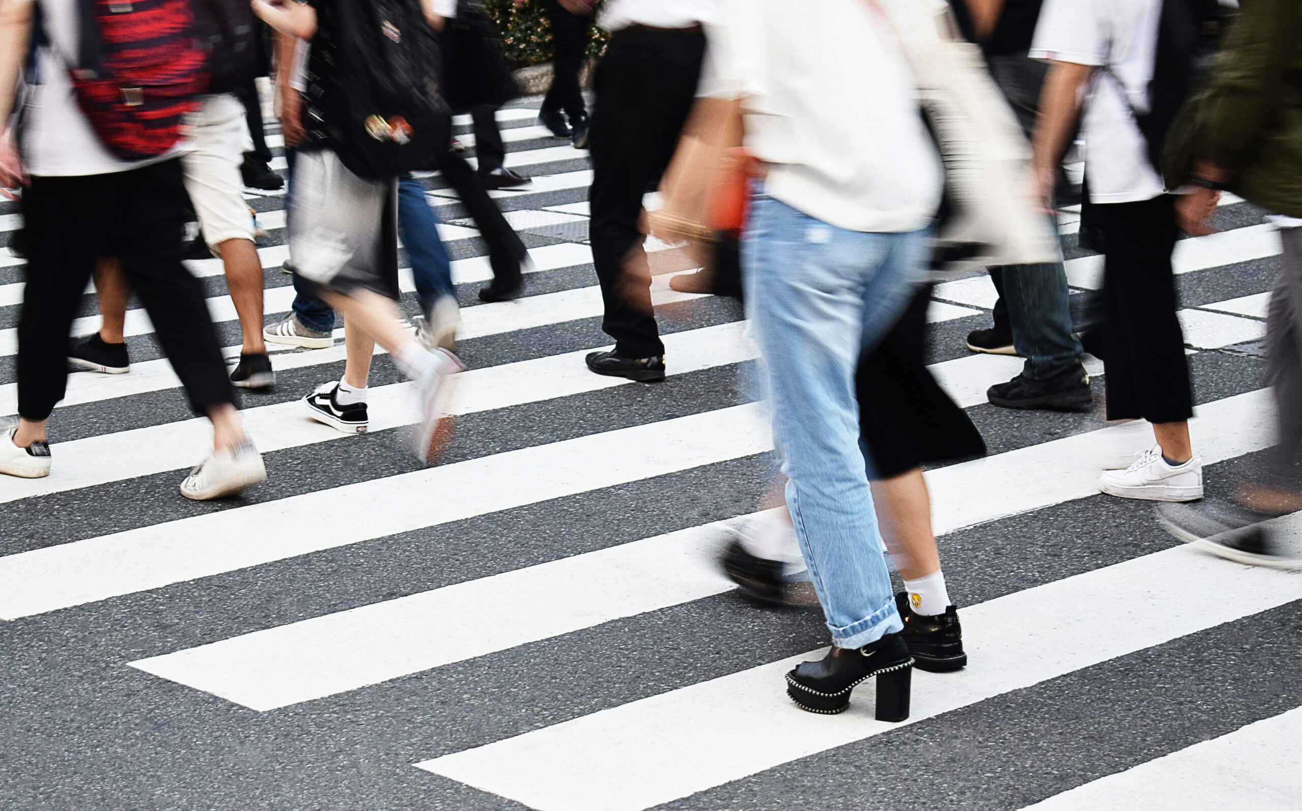Blurred motion of pedestrians crossing a zebra crosswalk, showing legs and feet moving across a busy street