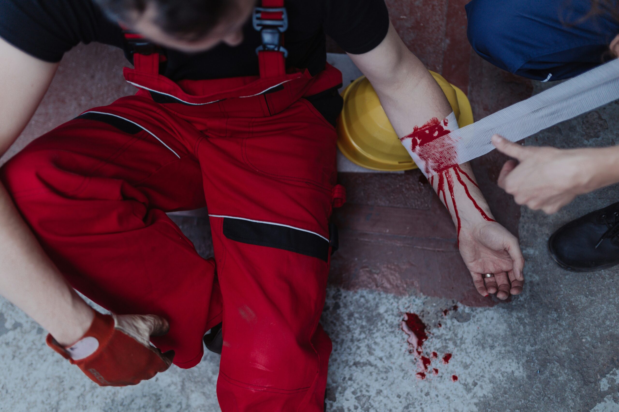 Worker in red overalls sitting on the ground with an injured arm being wrapped in a bandage, visible bleeding and a hard hat nearby