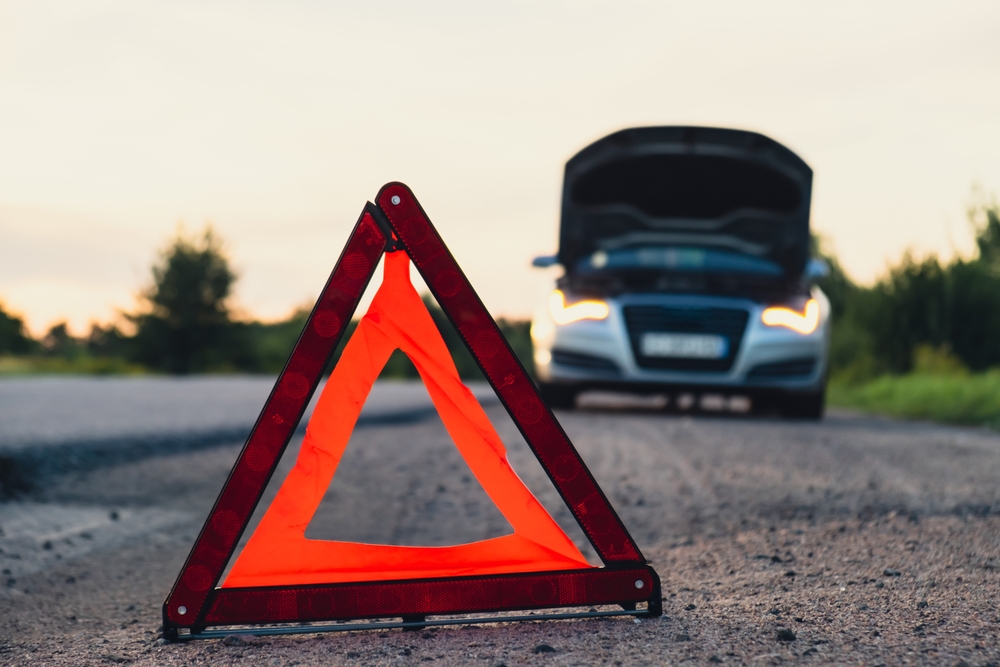 Reflective roadside warning triangle placed on a road in front of a disabled vehicle with its hood open in the background