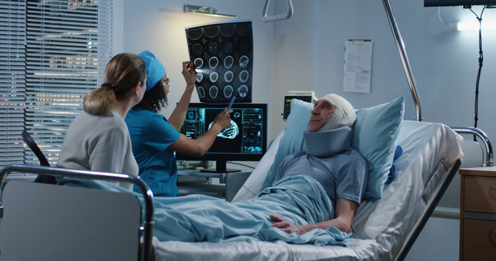 Nurse and a woman reviewing brain scans on a monitor beside a patient with a bandaged head and neck brace in a hospital bed