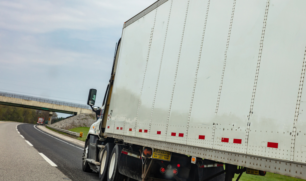 Semi truck with a box trailer driving on a highway near an overpass, with a sign on the back reading Caution: This vehicle makes wide left turns